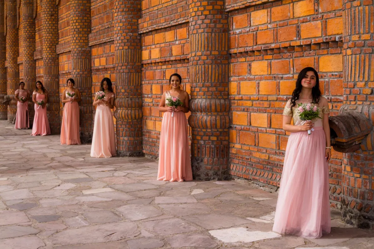 Bridesmaids in long pink dresses holding bouquets along a brick wall