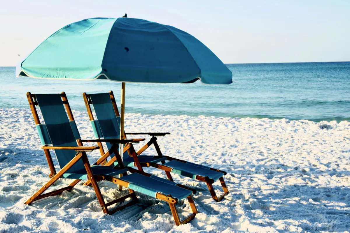 Two wooden folding beach chairs under a blue umbrella on white sand