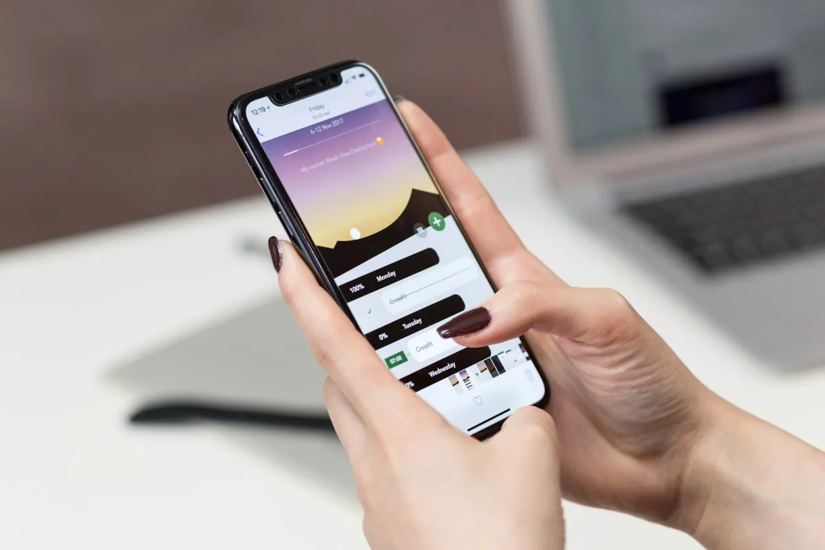 Hands holding a smartphone over a desk with a laptop in the background Hands holding a smartphone over a desk with a laptop in the background