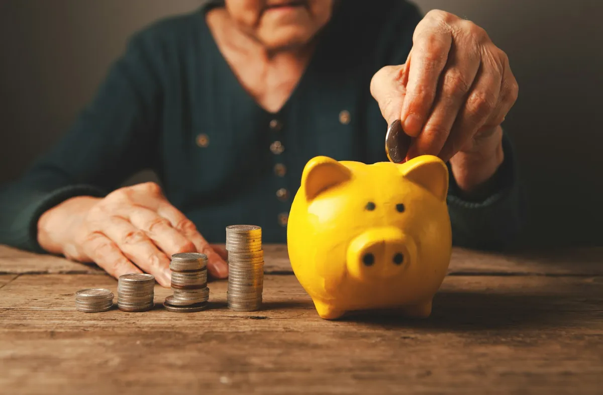 Elderly hands dropping a coin into a yellow piggy bank beside stacks of coins on a wooden table Elderly hands dropping a coin into a yellow piggy bank beside stacks of coins on a wooden table