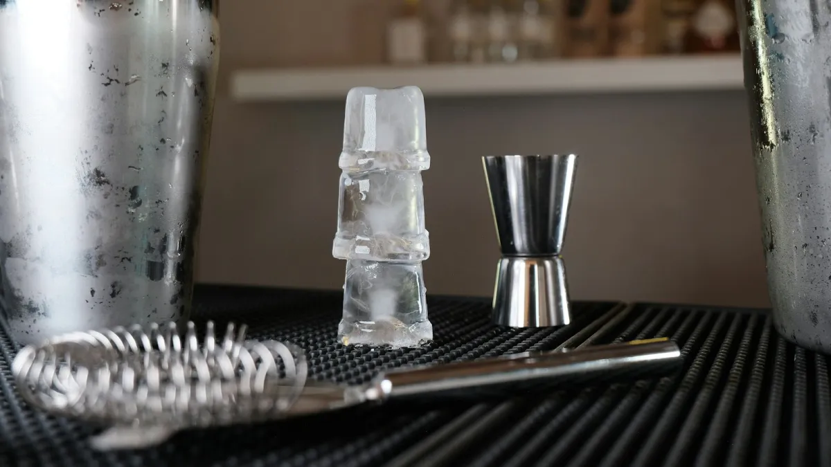 A cocktail shaker, jigger, strainer and stacked ice cubes on a bar mat A cocktail shaker, jigger, strainer and stacked ice cubes on a bar mat