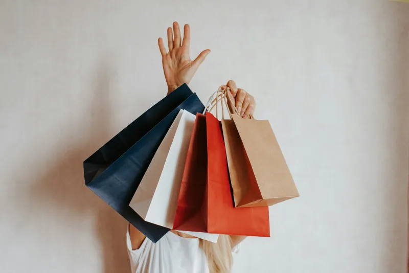 A person holding up several colorful paper shopping bags against a white wall