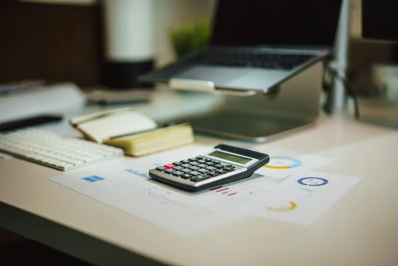 A calculator on printed charts on a desk, with a laptop and keyboard behind it