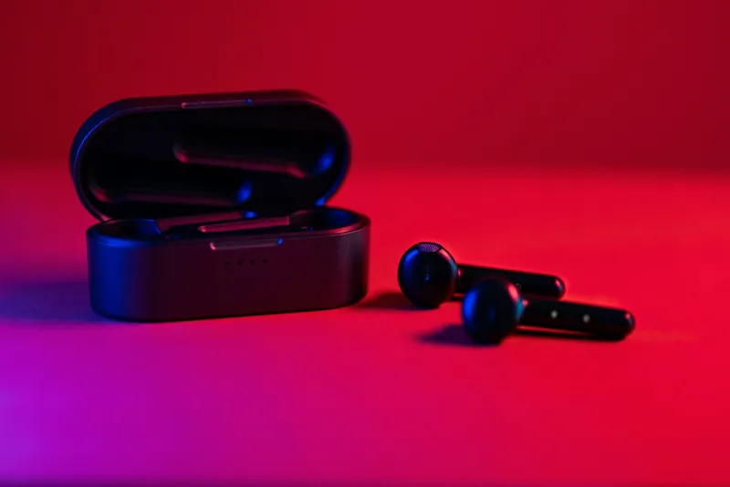 A pair of black wireless earbuds beside an open charging case under red and blue light