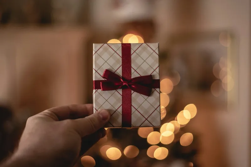 A hand holding a small gift box tied with a red ribbon in front of blurred holiday lights