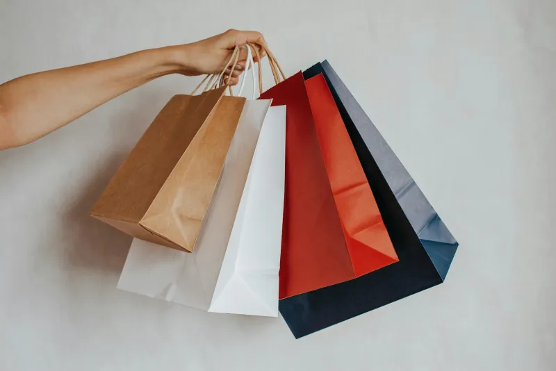 A hand holding several plain paper shopping bags in brown, white, red and navy against a light wall