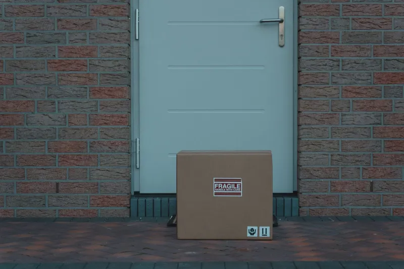 A cardboard box marked fragile left on the ground outside a closed front door in a brick wall