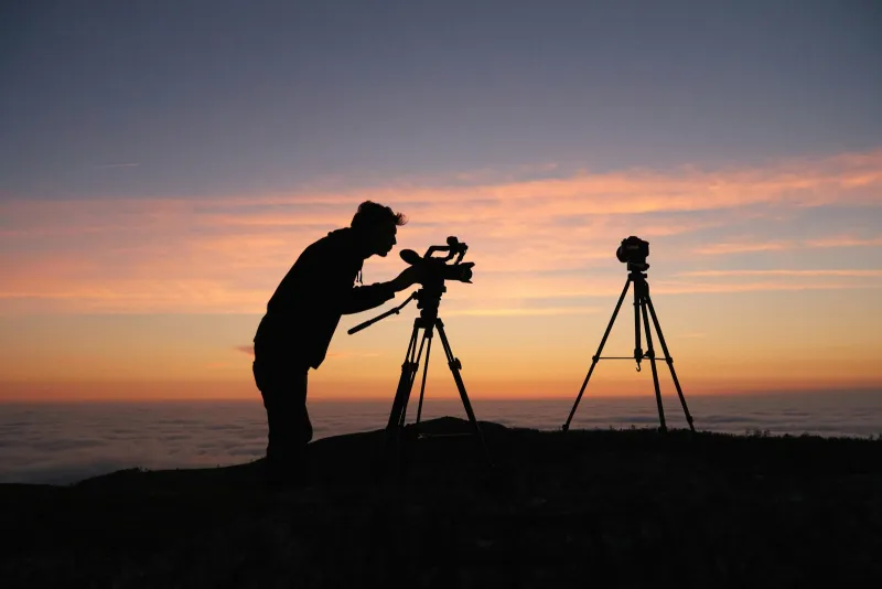 Silhouette of a videographer with two cameras on tripods against a sunset above the clouds