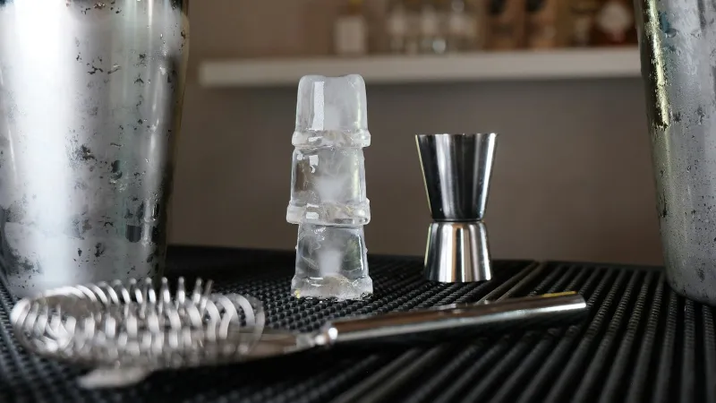 A cocktail shaker, jigger, strainer and stacked ice cubes on a bar mat